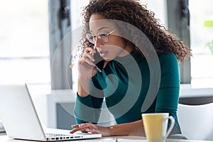 Confident young business woman talking on mobile phone while working with her laptop in the office