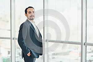 Confident young businessman standing, keeping hands in pockets, looking at the camera, ready to hard work day.