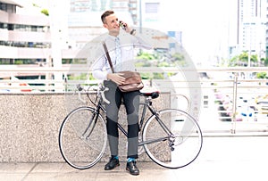 Confident young businessman using phone, looking away and smiling on his bike, standing city.