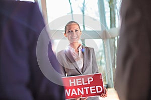 Confident young businesswoman holding red help wanted sign while standing at office