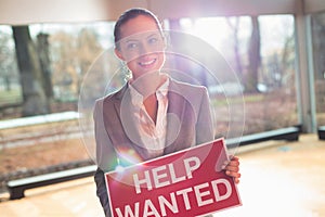 Confident young businesswoman holding red help wanted sign while standing at office