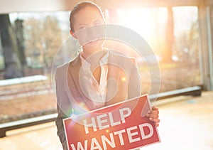 Confident young businesswoman holding red help wanted sign while standing at office