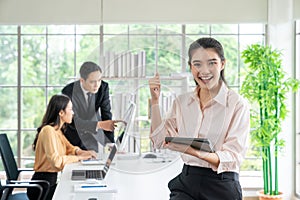 A confident young businesswoman holding tablet and looking at camera while her colleagues working in office background on business