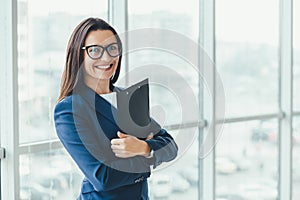 Confident young businesswoman standing with folder in hands, looking at the camera, ready to hard work day.