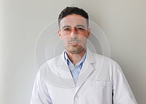Confident young Caucasian male doctor with dark brown hair in white hospital gown looking at camera on grey background, authentic