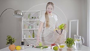 Confident young Caucasian woman taking out fruits and vegetables from bag at home. Pretty slim girl cooking healthful