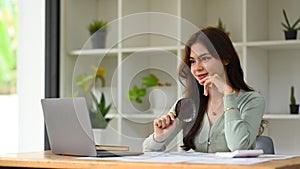 Confident young female auditor inspecting financial document with a magnifying glass at her office desk