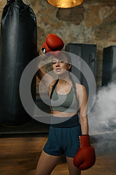 Confident young female boxer wearing gloves rest nearby punching bag