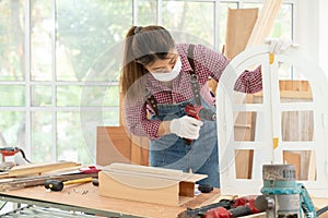 A confident young female carpenter uses a hand drill to assemble the wood window in the carpentry shop