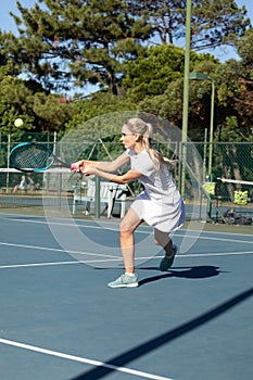 Confident young female caucasian tennis player hitting ball with racket at court on sunny day