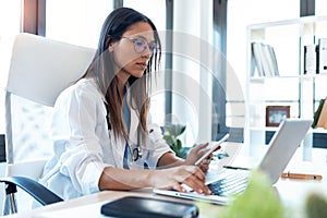 Confident young female doctor using her mobile phone while working on laptop in the consultation