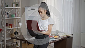 Confident young female freelancer standing in home office leaning on table typing on laptop keyboard. Portrait of