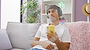 Confident, young hispanic man, chilling at home, sitting on his comfy sofa, enjoying his favorite tunes on his mobile by listening