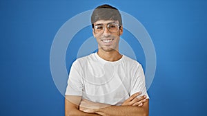 Confident young hispanic man wearing glasses and looking cool, standing with a cheerful smile and positive expression against an