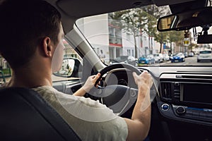 Confident young man driving car on the city streets. Teen driver keeps hands on the steering wheel looking ahead at the urban