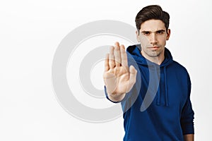 Confident young man extends hand to say stop, warning gesture, prohibit action, telling no, standing over white