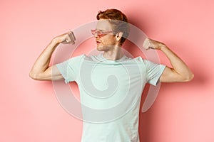 Confident young man with red hair, wearing summer sunglasses and t-shirt, showing strong and fit body muscles, flex