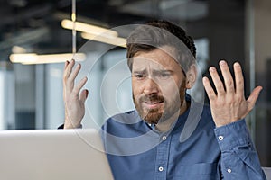 A confident young man sits in a modern office and looks disappointedly at the laptop screen, spreading his hands. Close