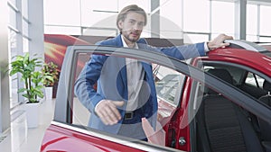 Confident young man standing at open red car in dealership, looking at camera and showing thumb up in slowmo. Portrait