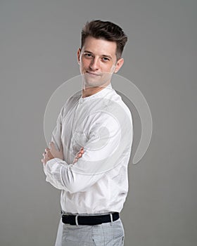 Confident young man standing in white shirt on grey background. Body profile with crosed hands