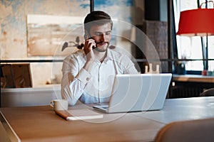 Confident young man working on laptop and talking on the mobile phone while sitting at his working place in office