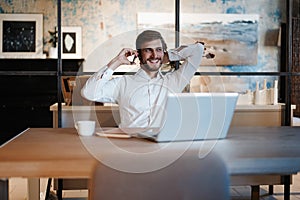 Confident young man working on laptop and talking on the mobile phone while sitting at his working place in office