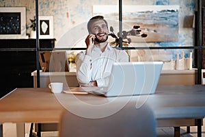 Confident young man working on laptop and talking on the mobile phone while sitting at his working place in office