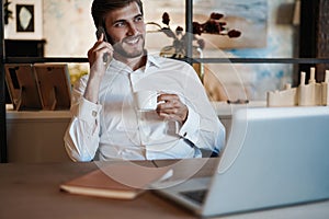 Confident young man working on laptop and talking on the mobile phone while sitting at his working place in office