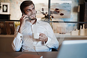 Confident young man working on laptop and talking on the mobile phone while sitting at his working place in office