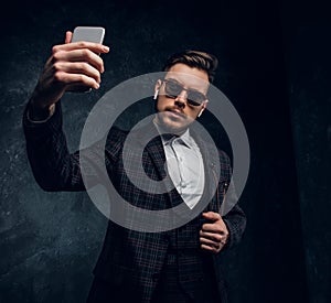 Confident young stylish man dressed an elegant suit taking selfie by smartphone in a dark studio