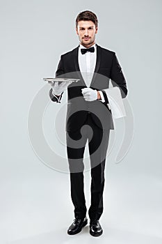 Confident young waiter in tuxedo standing and holding tray