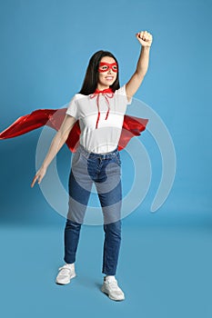 Confident young woman wearing superhero cape and mask on light blue background