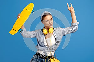 Confident young woman with yellow headphones posing with skateboard showing rock gesture over blue background.
