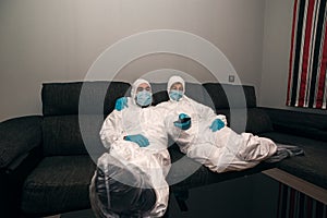 Confined couple embracing each other during curfew watching TV wearing nbc protective suits against the covid-19 coronavirus