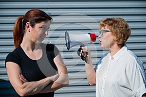 The conflict of generations. An old woman yells into a megaphone at her middle-aged daughter against gray background