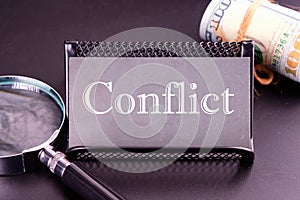 Conflict word on the business card next to a roll of money and a magnifying glass on a black background