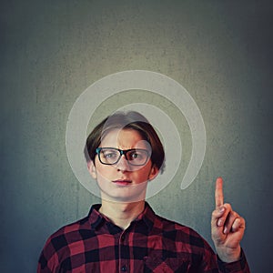 Confused adolescent guy, wearing eyeglasses gesturing, pointing index finger up, perplexed expression isolated on grey wall
