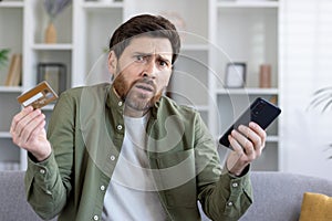 Confused adult man holding smartphone and bank card in hands while shrugging with worried expression. Uncertain customer