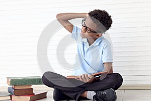 Confused African boy with glasses reading book while sitting on floor in white wall room. Stressed child with pile of books try to