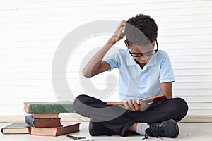 Confused African boy with glasses reading book while sitting on floor in white wall room. Stressed child with pile of books try to