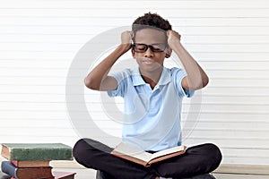 Confused African boy with glasses reading book while sitting on floor in white wall room. Stressed child with pile of books try to