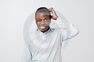 Confused african man scratching head while looking at camera and standing over gray background.