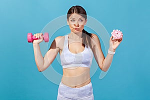 Confused athletic woman in white sportswear holding in hands pink dumbbell and donut with pink glaze, making hard choice, succumb photo