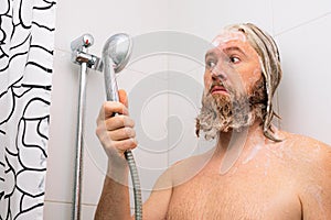 Confused bearded man with soapy head standing in bathroom and looking at the shower while the water supply has stopped