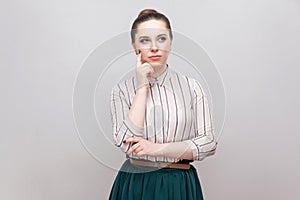 Confused beautiful young woman in striped shirt and green skirt with makeup and collected ban hairstyle, standing thoughtful and