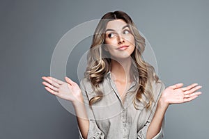 Confused blonde woman with curly hair dressed in linen shirt spreads hands sideways and shrug shoulders, smirks face, looking up