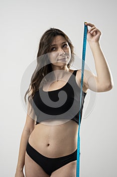 Confused brunette female with perfect slim body in a black underwear looking on a blue measure tape. Healthy nutrition