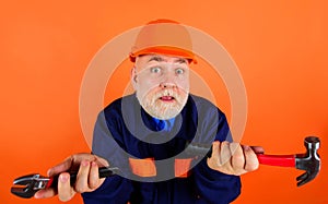 Confused builder in safety hard hat with adjustable wrench and hammer. Bearded mechanical worker or repairman in uniform