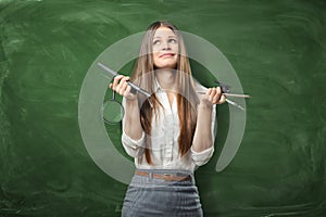 Confused businesswoman on green chalkboard background holding pens, ruler and magnifying glass in her hands.