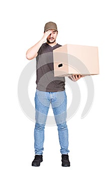 Confused delivery man, full length portrait, holding a cardboard box while keeps a hand to forehead, isolated on white. Puzzled